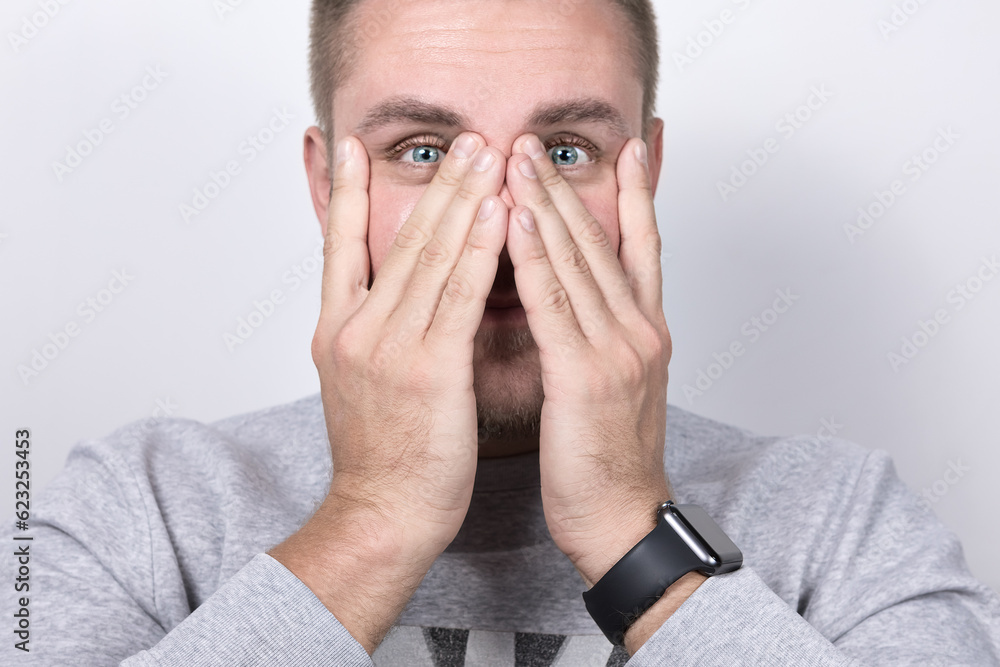 Vision problems, myopia and astigmatism,sight correction. A young man with blue eyes tries to focus his vision by pulling on the skin and muscles around his eyes, his face half covered by his hands Vision problems, myopia and astigmatism,sight correction. A young man with blue eyes tries to focus his vision by pulling on the skin and muscles around his eyes, his face half covered by his hands