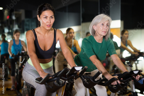 Young and adult confident female warming up on bikes in cycling class at gym