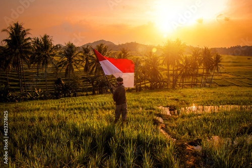 Photo of a village boy waving the Indonesian flag in the middle of a rice field with the silhouette of an optical sunset flair