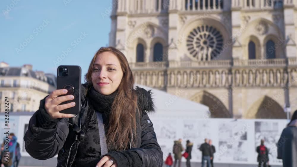 Positive Latina woman tourist smiling and taking selfie on smartphone while standing against landmark of Paris, France in Notre Dame cathedral Positive Latina woman tourist smiling and taking selfie on smartphone while standing against landmark of Paris, France in Notre Dame cathedral