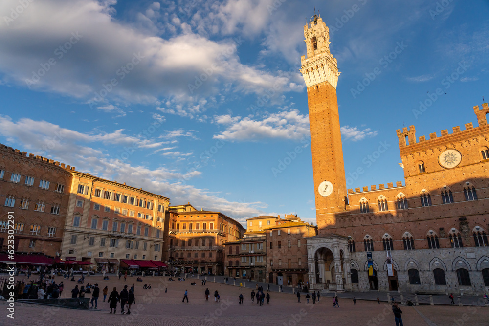 Sunset in the Piazza del Campo square of Siena, Italy. The Torre del Mangia is bathed in golden light, as well as the tops of some buildings. People visible on the town square.