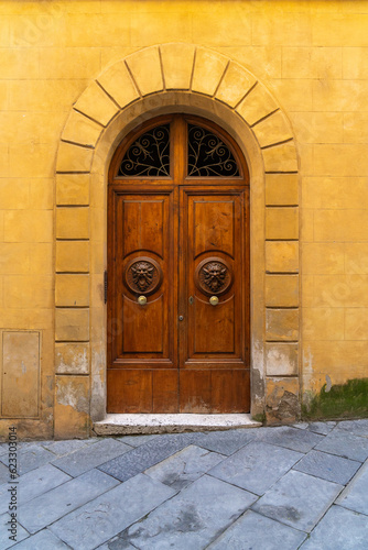 Medieval brown wooden door with two lion head doorknobs set inside a buttercream yellow colored arched doorway and wall in the city of Siena, Italy.  © Patrick