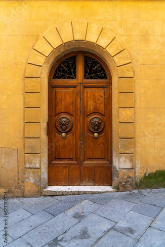 Medieval brown wooden door with two lion head doorknobs set inside a buttercream yellow colored arched doorway and wall in the city of Siena  Italy. 