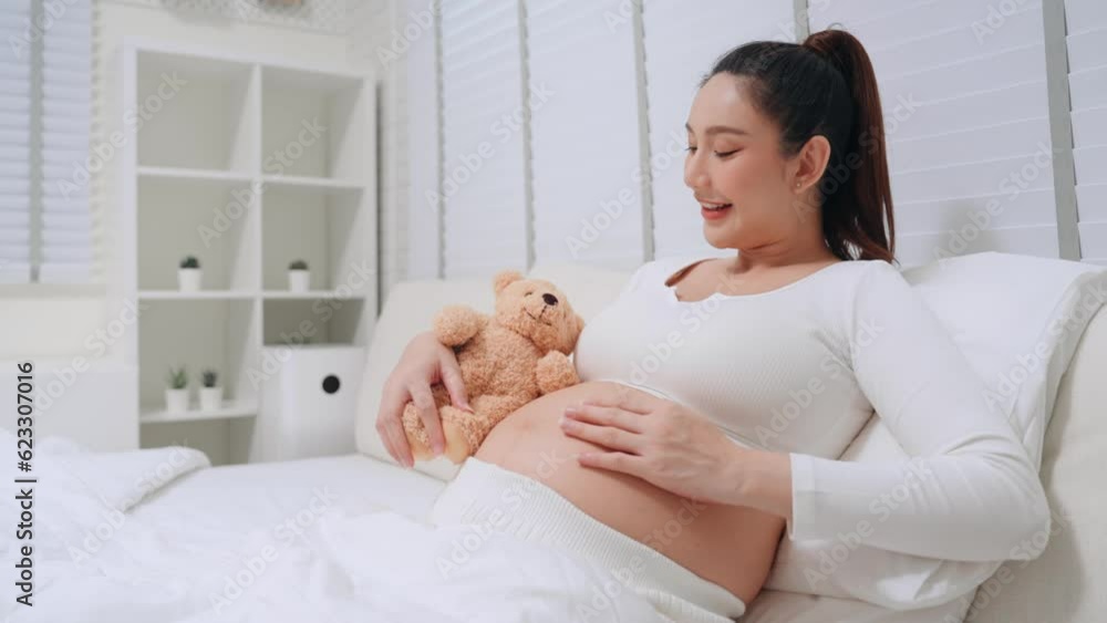 Delight pregnant asian woman stroking and touching belly slowly with love and play brown bear doll talking with baby in tummy on bed at home. Asian mother happiness pending baby in tummy prenatal. Delight pregnant asian woman stroking and touching belly slowly with love and play brown bear doll talking with baby in tummy on bed at home. Asian mother happiness pending baby in tummy prenatal.