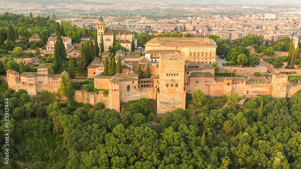 Aerial sunrise image of the Alhambra fortress in Granada, Spain. The fortress is bathed in reddish-orange light with green trees interspersed among towers.