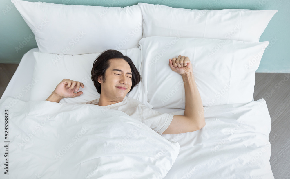 © paulaphoto - Portrait of handsome young asian man with enjoy fresh soft bedding linen mattress in white bed room modern retro apartment. Asian guy sleep stretch resting, good night sleep concept banner