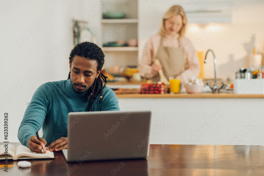 A multicultural casual entrepreneur is sitting at a table with a laptop and working while his wife is cooking in the kitchen. A multicultural casual entrepreneur is sitting at a table with a laptop and working while his wife is cooking in the kitchen.
