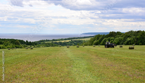 agricultural field with bales of hay and working tractor with forest line and river  