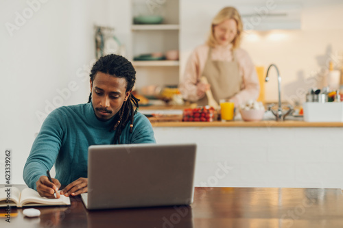 A multicultural casual entrepreneur is sitting at a table with a laptop and working while his wife is cooking in the kitchen. © Zamrznuti tonovi