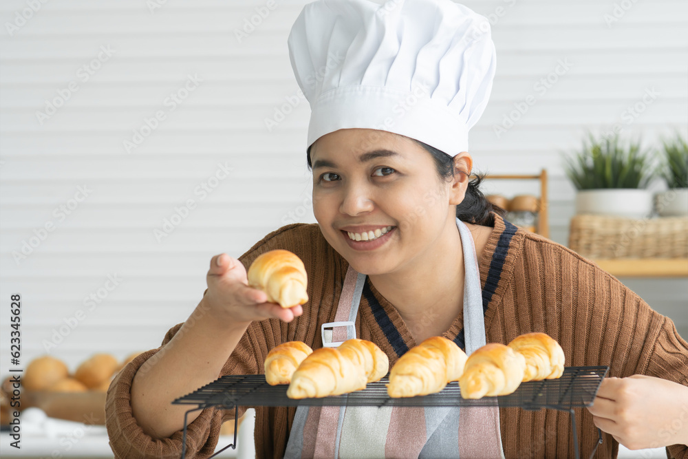 Portrait of smiling woman in apron and chef hat baking pastry at home kitchen, looking at camera, checking delicious baked croissants out of oven. Joyful homemade baker showing bread in hand Portrait of smiling woman in apron and chef hat baking pastry at home kitchen, looking at camera, checking delicious baked croissants out of oven. Joyful homemade baker showing bread in hand