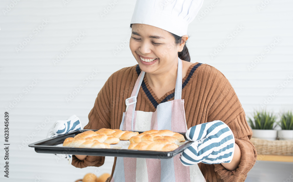 Portrait of smiling woman in apron and chef hat baking pastry at home kitchen, looking at delicious baked breads out of oven with pleasure. Joyful homemade baker holding tray in hands with gloves Portrait of smiling woman in apron and chef hat baking pastry at home kitchen, looking at delicious baked breads out of oven with pleasure. Joyful homemade baker holding tray in hands with gloves