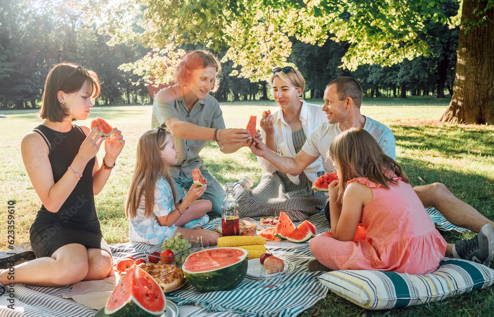 Big family sitting on the picnic blanket in a city park during weekend Sunday sunny day. They are smiling, chatting, laughing and eating juicy watermelon. Family values and outdoor activities concept. Big family sitting on the picnic blanket in a city park during weekend Sunday sunny day. They are smiling, chatting, laughing and eating juicy watermelon. Family values and outdoor activities concept.