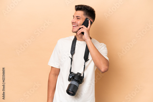 Young photographer caucasian man isolated on beige background keeping a conversation with the mobile phone © luismolinero