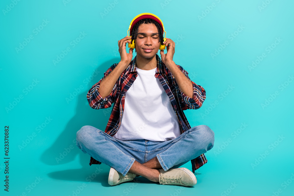 Full length photo of peaceful satisfied young man sitting floor closed eyes arms touch headphones isolated on teal color background Full length photo of peaceful satisfied young man sitting floor closed eyes arms touch headphones isolated on teal color background