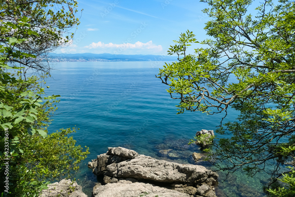 A panoramic view of the Croatian shore. Big rocks in the water, shore is overgrown with lust, green plants. There are some towns located on the shore of the Mediterranean Sea. Green hills in the back
