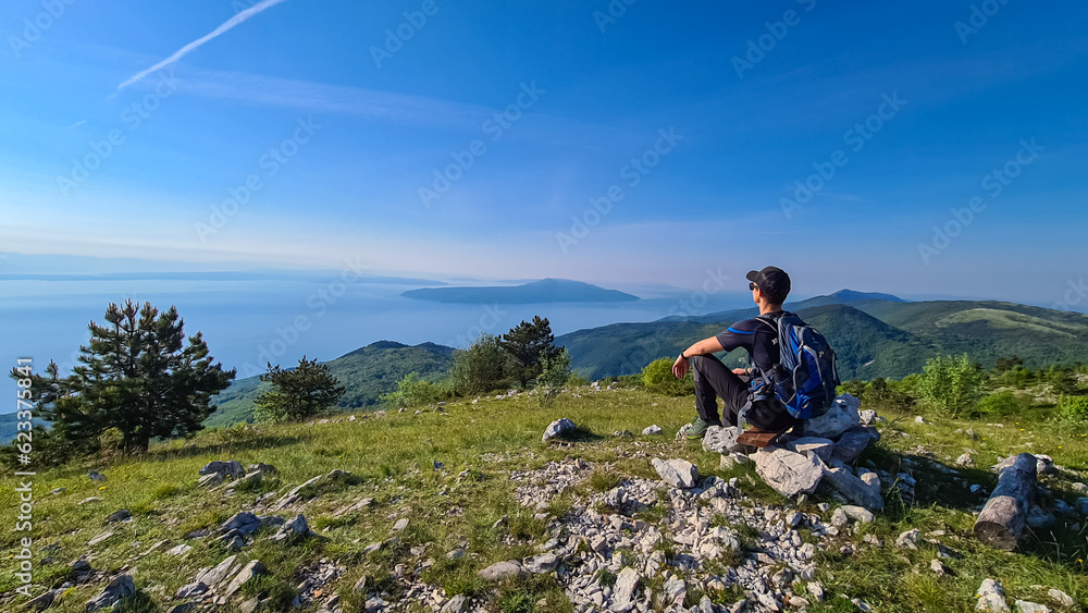 Obraz premium A man sitting on top of Vojak and enjoying the panoramic view on Mediterranean Sea. The mountain is overgrown with lush green plants. Few islands in the back. Early morning hiking by the sea. Remedy