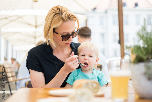 Young caucasian blonde mother spoon feeding her little infant baby boy child outdoors on restaurant or cafe terrace in summer.
