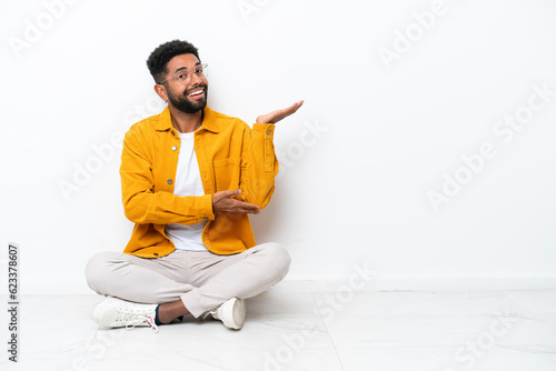 Young Brazilian man sitting on the floor isolated on white background extending hands to the side for inviting to come