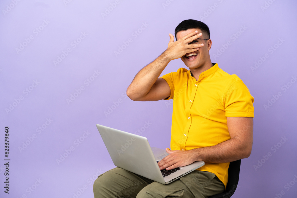 Young man sitting on a chair with laptop covering eyes by hands and smiling Young man sitting on a chair with laptop covering eyes by hands and smiling