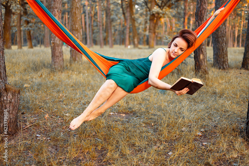 Young beautiful blonde girl in hammock reading book. Pretty woman leisure lifestyle portrait at nature ountdoor. Female relax in forest. Tourism in summer camp. Hobby photo