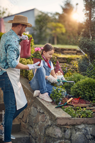 Girl sitting by the outdoor garden assiting her father in gardening, planting together, young man holding watering can and flower ready to plant.