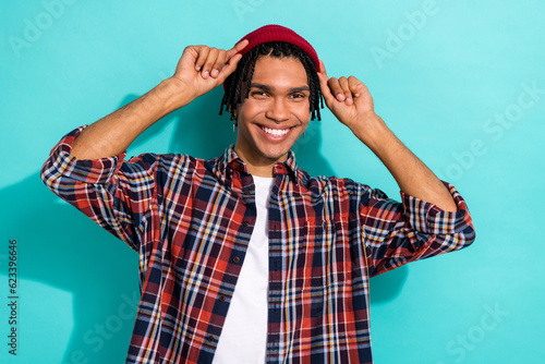 Photo of satisfied cool nice guy with cornrows dressed checkered shirt arms touching stylish hat isolated on teal color background