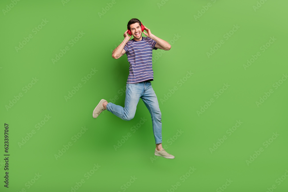 Full length photo of cheerful funky man dressed striped t-shirt jumping high enjoying songs isolated green color background