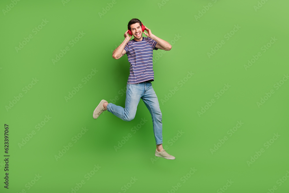 Full length photo of cheerful funky man dressed striped t-shirt jumping high enjoying songs isolated green color background Full length photo of cheerful funky man dressed striped t-shirt jumping high enjoying songs isolated green color background