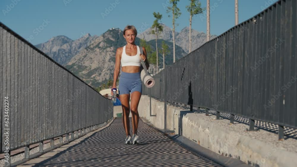 Athlete girl walking seacoast at twilight holding sport mat. Yoga woman going workout wearing sportswear outdoors. Attractive confident sportswoman enjoying training in beautiful summer evening Athlete girl walking seacoast at twilight holding sport mat. Yoga woman going workout wearing sportswear outdoors. Attractive confident sportswoman enjoying training in beautiful summer evening