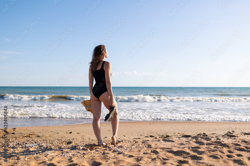Beautiful brunette woman in a black bodysuit is resting in the summer on a sandy seashore under a blue sky