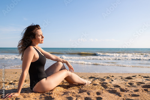 Beautiful brunette woman in a black bodysuit is resting in the summer on a sandy seashore under a blue sky