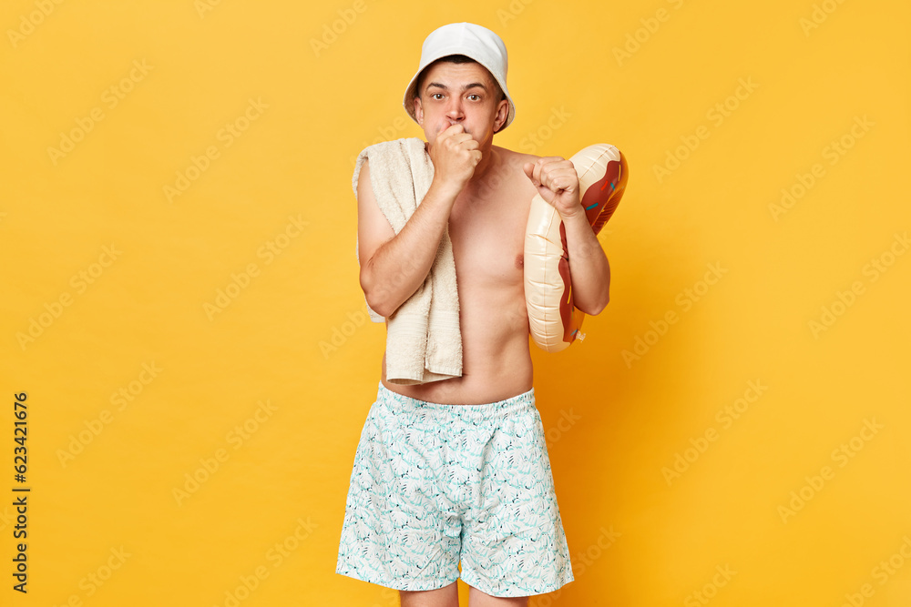 Funny confused man man wearing shorts swimsuit and panama holding donut rubber ring isolated on yellow background clenched fist looking at camera. Funny confused man man wearing shorts swimsuit and panama holding donut rubber ring isolated on yellow background clenched fist looking at camera.