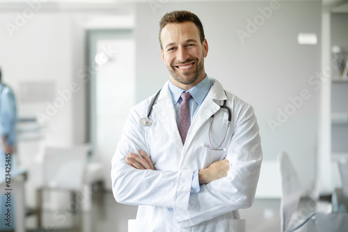 Happy healthcare worker. Portrait of confident doctor in white uniform with stethoscope posing with folded arms smiling at camera at medical clinic