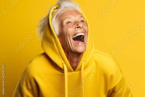 Headshot portrait photography of a joyful old woman wearing a comfortable hoodie against a bright yellow background. With generative AI technology