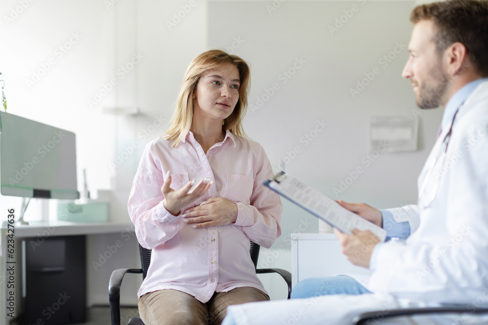 Obraz premium Checkup during pregnancy. Young european expecting woman visiting doctor man and talking with therapist in private clinic