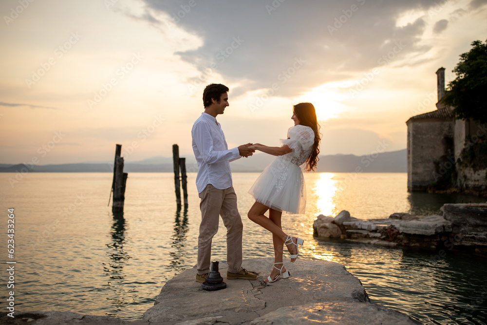 Fototapeta premium Romantic just married couple dancing looking to each other at a medieval stone pier of Italian village at Lago di Garda at the golden sunset