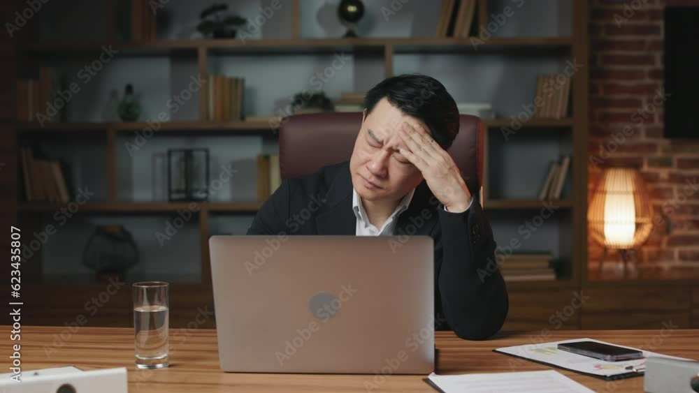 Overworked asian employee suffering from strong migraine and touching head with closed eyes. Exhausted man massaging temples in front of laptop in order to relief emotional stress and disorder. Overworked asian employee suffering from strong migraine and touching head with closed eyes. Exhausted man massaging temples in front of laptop in order to relief emotional stress and disorder.