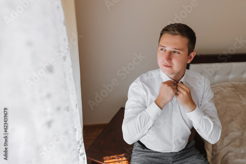 attractive young man poses as a groom before the wedding, adjusting the collar of his shirt. the groom is preparing for the wedding in a large building