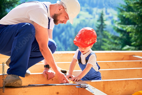 Father with toddler son building wooden frame house. Male worker teaching his son how to measure angle on construction site, wearing helmets and overalls on sunny day. Carpentry and family concept. photo