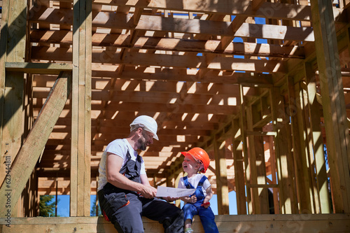 Father with toddler son building wooden frame house. Boys having fun on the edge of balcony, examining the construction plan, wearing helmets and overalls on sunny day. Carpentry and family concept.