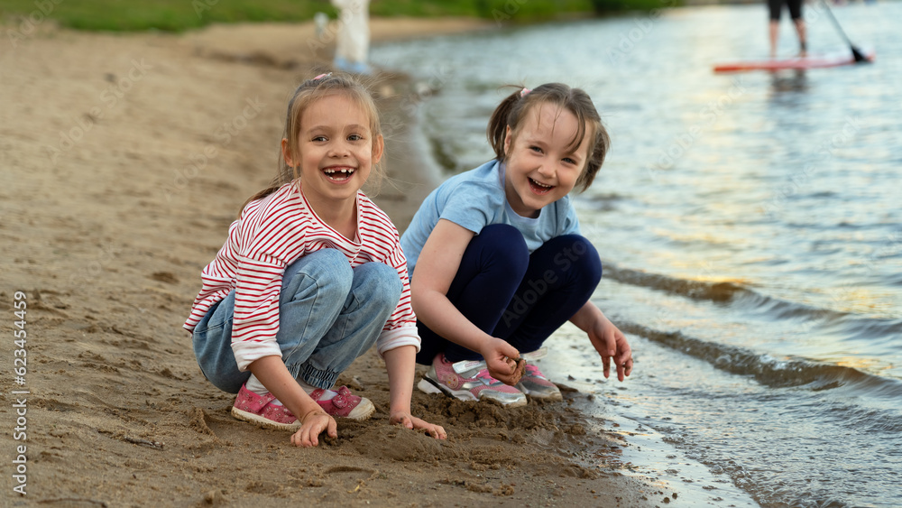 Happy little girls look at camera and laugh on sand river bank in summer park at sunset. Children walk outdoors by water Happy little girls look at camera and laugh on sand river bank in summer park at sunset. Children walk outdoors by water