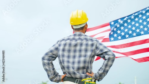 Confidently standing construction working with safety hat in front of waving American Flag - concept of Independence day or Labours day celebration