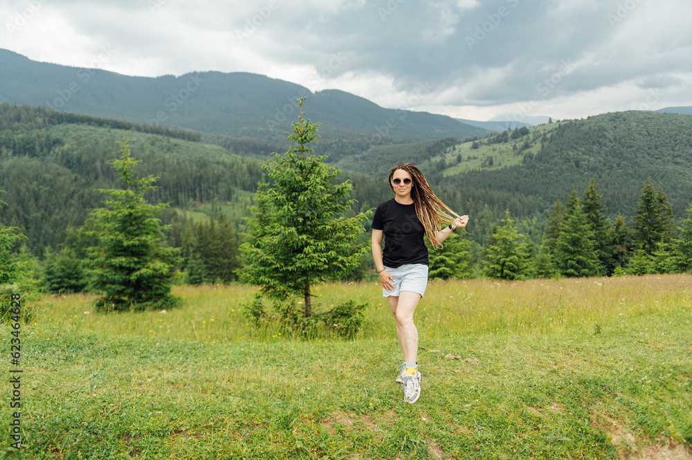 Obraz premium A beautiful woman with a hairstyle - a lot of braids, posing at the top of the mountain in a black T -shirt and shorts.