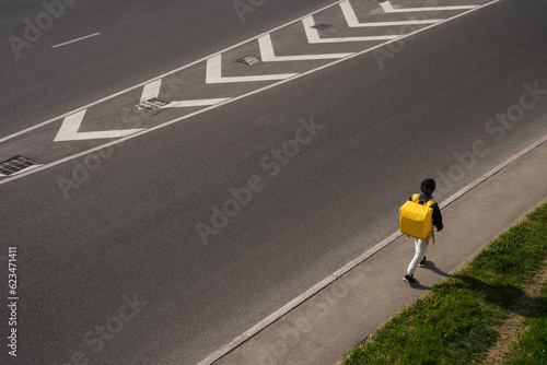 A man walks along the sidewalk near the road with a yellow delivery bag