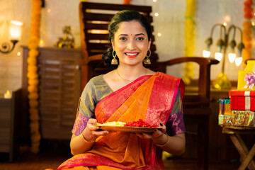 Indian happy smiling girl holding flower plate for diwali celebration by looking at camera at home concept of - indian traditional, ceremony and festival preparation.