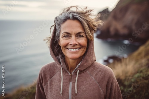 Close-up portrait photography of a happy mature woman wearing a comfortable hoodie against a dramatic coastal cliff background. With generative AI technology