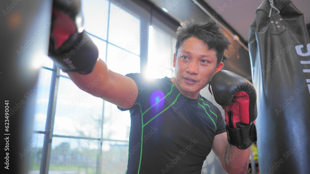 Thai boxing man in gym with sandbag. Athlete man in fitness with glove train in punching and kicking. Martial art exercise for strength.