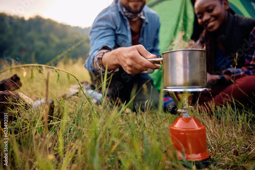 Close up of multiracial couple making tea while using portable camping stove in nature.