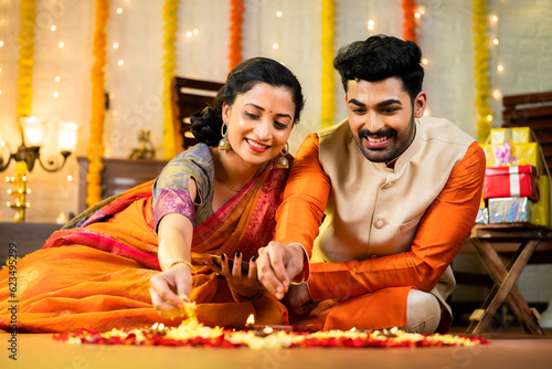 Happy indian couple in traditional ethnic wear decorating rangoli with flower for festival celebration while sitting on floor at home concept of - relationship, bonding, and religious ceremony.