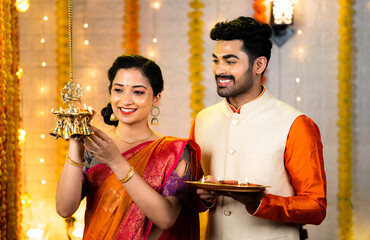 Happy indian couple in traditional ethnic clothing lighting brass hanging oil lamp during diwali festival celebration at home - concept of diwali celebration, auspicious and indian culture